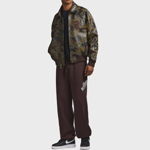 May include: A camouflage-patterned jacket with a brown ribbed hem and cuffs. The jacket has a brown patch on the chest. The person is wearing brown sweatpants with a white logo on the left leg and black sneakers.