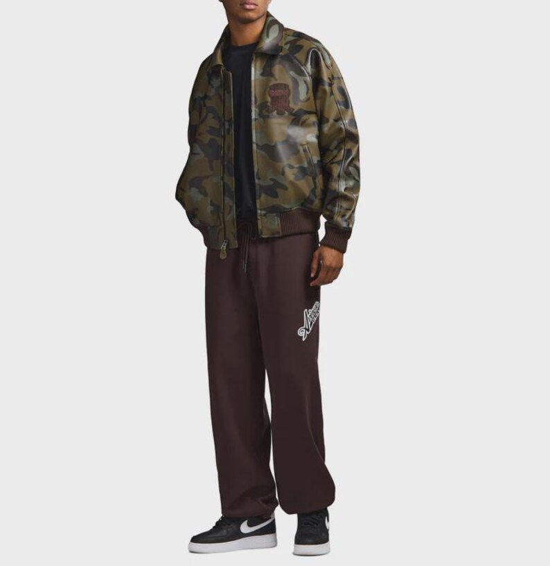 May include: A camouflage-patterned jacket with a brown ribbed hem and cuffs. The jacket has a brown patch on the chest. The person is wearing brown sweatpants with a white logo on the left leg and black sneakers.