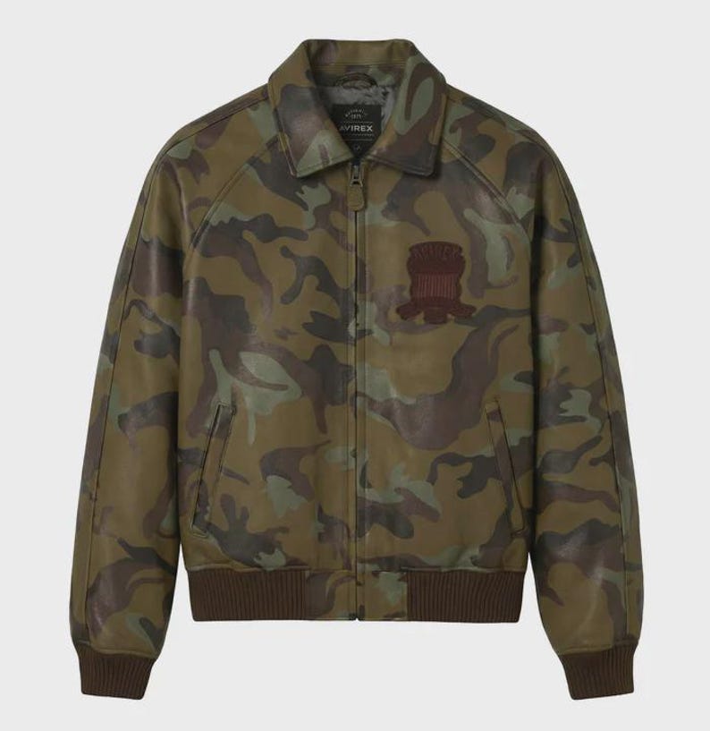 May include: A camouflage-patterned leather jacket in shades of green, brown, and tan. The jacket features a collared neckline, a zipper closure, and a brown ribbed hem and cuffs. A brown patch is on the chest. The brand name "AVIREX" is visible on the collar.