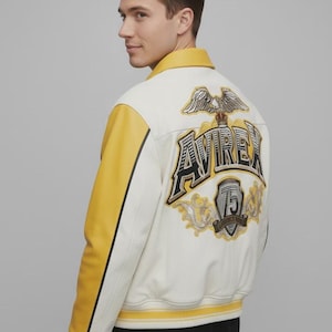 May include: Cream-colored leather jacket with yellow sleeves and collar. The back features an embroidered eagle and the word "AVIREX" in a bold, stylized font. A "75" emblem is also embroidered on the back. The jacket has a classic bomber style.