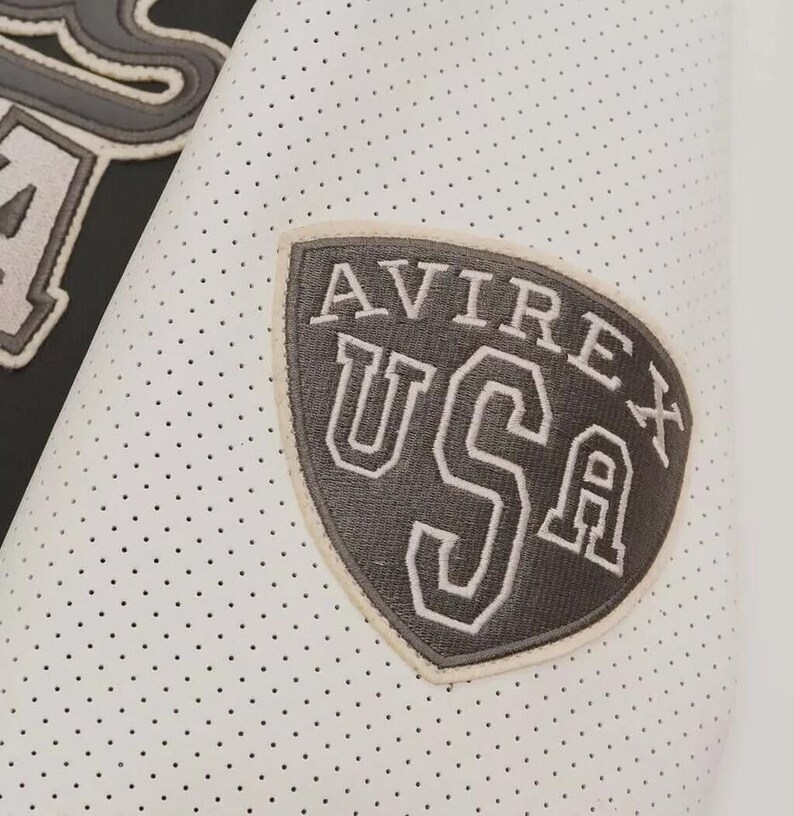May include: Close-up of a white perforated fabric with a gray and white patch. The patch reads "AVIREX USA" in a shield shape. The fabric has small, evenly spaced holes.