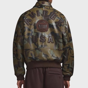May include: Camouflage-patterned bomber jacket with brown ribbed cuffs and hem. The back features the word "USA" and the brand name "AVIREX" in brown lettering. A brown emblem is centered above the "USA" text.