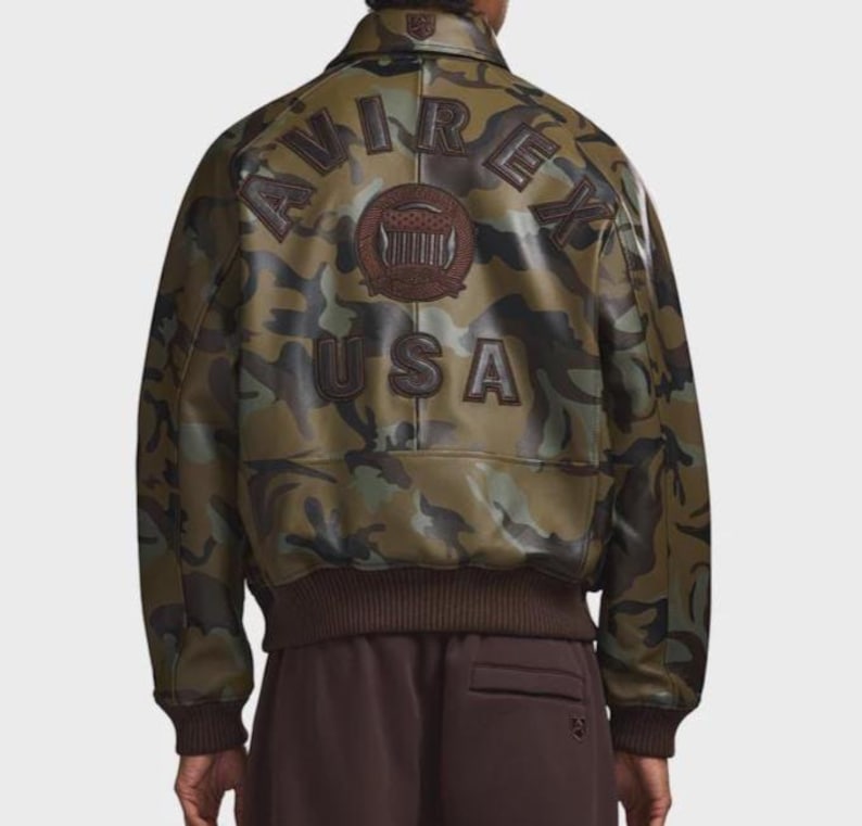 May include: Camouflage-patterned bomber jacket with brown ribbed cuffs and hem. The back features the word "USA" and the brand name "AVIREX" in brown lettering. A brown emblem is centered above the "USA" text.