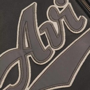 May include: Close-up of a black leather item with the word "Air" stitched in gray and outlined in white. The stitching is detailed, and the leather has a subtle texture. The zipper is visible on the right side.