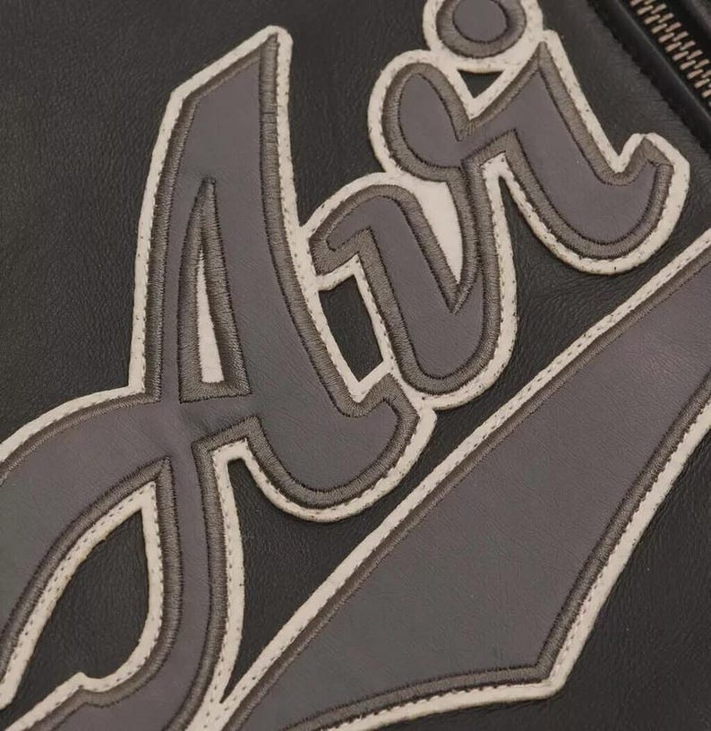 May include: Close-up of a black leather item with the word "Air" stitched in gray and outlined in white. The stitching is detailed, and the leather has a subtle texture. The zipper is visible on the right side.
