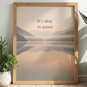 May include: A framed print with a tranquil mountain and lake scene, and the words "It's okay to pause". The frame is light wood. Potted plants flank the frame. The image evokes a sense of calm and reflection.
