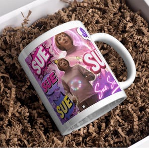 May include: A white ceramic mug featuring a repeating graphic design of a person with short brown hair wearing a light-coloured turtleneck. The design includes the name SUE in various colourful comic-style speech bubbles and neon pink text.