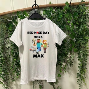May include: White t-shirt with a graphic featuring the text "RED NOSE DAY 2026" above a pixelated illustration of characters and a dog. The name "MAX" is printed below the graphic. The shirt is hanging on a black hanger.