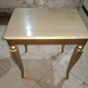 Op de afbeelding: Een kleine, vierkante bijzettafel met een lichtbeige blad en goudkleurige poten. De tafel heeft een glanzende afwerking en een klassiek ontwerp, geschikt voor diverse interieurstijlen. De poten zijn gebogen en voegen een vleugje elegantie toe.