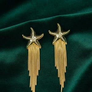 Può includere: Un paio di orecchini a forma di stella marina color oro con lunghe catene pendenti. Gli orecchini sono posti su uno sfondo verde scuro strutturato. Il design a stella marina è dettagliato e tridimensionale.