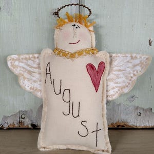 May include: A fabric angel ornament in cream with yellow hair, a gold wire halo, and white wings. The body features the embroidered text "August" in black thread and a red stitched heart. The angel has a simple hand-drawn face with pink cheeks.