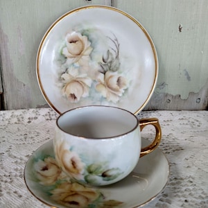 May include: A vintage porcelain teacup, saucer, and plate set. The white pieces feature hand-painted yellow roses with green leaves and gold trim. The teacup has a gold handle. The plate and saucer have a gold rim.