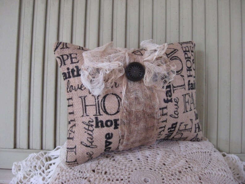 May include: A beige pillow with a lace trim and a button. The pillow is made of burlap and has the words "Hope", "Faith", and "Love" printed on it.