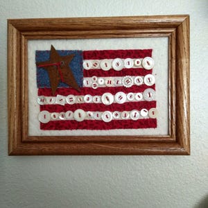 May include: Framed artwork depicting the American flag. The flag features red and white stripes made of fabric, with white buttons sewn on. The blue field has a rusty star. The artwork is framed in a wooden frame.