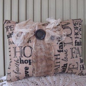 May include: A burlap pillow with a lace overlay and a brown button. The pillow is printed with the words "HOPE", "FAITH", "LOVE", and "ALL I HAVE IS FAITH".
