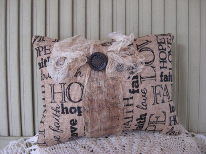 May include: A burlap pillow with a lace overlay and a brown button. The pillow is printed with the words "HOPE", "FAITH", "LOVE", and "ALL I HAVE IS FAITH".