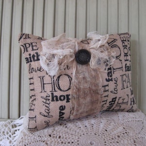 May include: A beige pillow with a lace trim and a button. The pillow is made of burlap and has the words "Hope", "Faith", and "Love" printed on it.