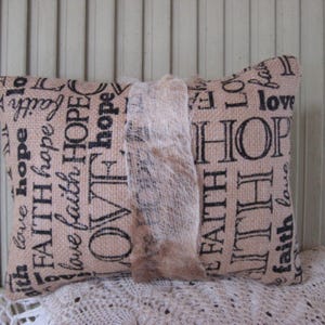 May include: A beige throw pillow with a burlap-like texture and black text that reads "with love hope FAITH hope love faith HOPE OVF hope FAITH AITH faith love love HOP lo LO fal".