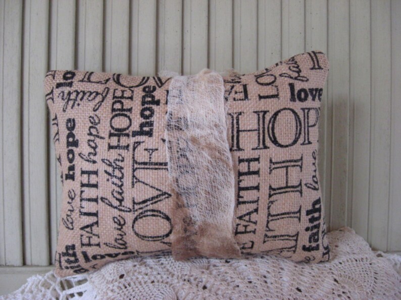 May include: A beige throw pillow with a burlap-like texture and black text that reads "with love hope FAITH hope love faith HOPE OVF hope FAITH AITH faith love love HOP lo LO fal".
