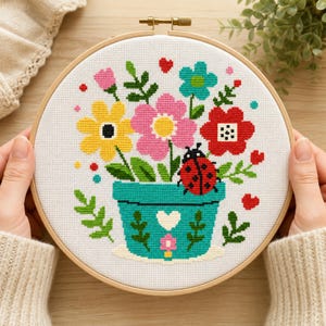 Könnte beinhalten: Ein Kreuzstich-Stickprojekt in einem runden Holzrahmen, das einen türkisfarbenen Blumentopf mit einem weißen Herz zeigt, gefüllt mit gelben, rosa, roten und türkisfarbenen Blumen, grünen Blättern und einem roten Marienkäfer mit schwarzen Punkten.