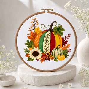 May include: A circular wooden embroidery hoop containing a cross-stitch pattern of a large, multi-coloured pumpkin surrounded by autumn leaves, a sunflower, and a smaller green and white striped pumpkin on a white fabric background.