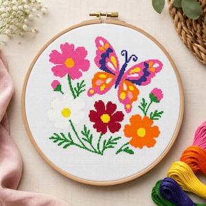 Op de afbeelding: Een ronde houten borduurring met witte stof voorzien van een kruissteekpatroon van een kleurrijke vlinder en diverse bloemen in roze, witte, rode en oranje tinten, met groene stengels en bladeren op de stof geborduurd.