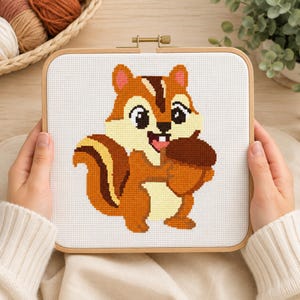 May include: A square wooden embroidery hoop containing white Aida fabric with a cross-stitch pattern of a brown and cream chipmunk holding a large brown acorn. The chipmunk has dark brown stripes on its head and tail, large black eyes, and a small smile.