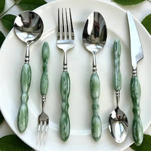 May include: A six-piece stainless steel flatware set with green and white marbled handles. The set includes a spoon, fork, knife, and two smaller spoons. The utensils are arranged on a white plate, showcasing a modern dining aesthetic.