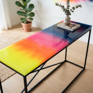 Könnte beinhalten: Ein rechteckiger Konsolentisch mit einer glatten, glänzenden Tischplatte aus Kunstharz in einem lebendigen Farbverlauf von Gelb, Orange, Pink und Dunkelblau. Der Tisch steht auf einem dünnen, schwarzen Metallrahmen mit Kreuzstreben.