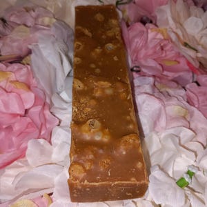 May include: A rectangular bar of brown soap with a textured surface. The soap has a light brown colour with small, lighter-coloured inclusions throughout. The soap is sitting on a bed of pink and white flowers.