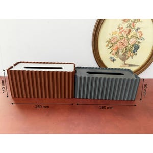 May include: Two rectangular tissue box covers with a vertical ribbed texture. The brown cover measures 250 mm by 110 mm, and the gray cover measures 250 mm by 90 mm. Both feature a central rectangular opening on the top for dispensing tissues.