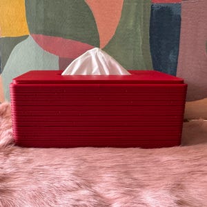 May include: A rectangular, deep red tissue box cover with a layered, ribbed texture on the exterior. The cover features a central rectangular opening at the top, through which a single white tissue is pulled upwards in a pointed, triangular shape.