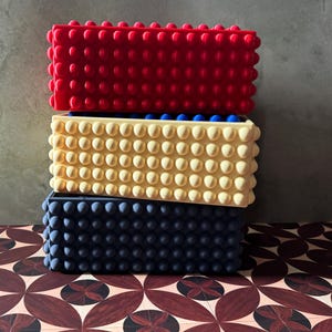 May include: A stack of three rectangular silicone molds with a textured surface featuring rows of raised circular bumps. The top mold is bright red, the middle mold is pale yellow with blue interior details, and the bottom mold is dark navy blue.