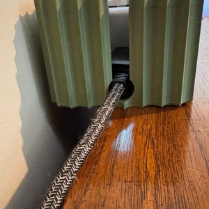 May include: A sage green, 3D-printed cable management box with a vertical ribbed texture. The rectangular enclosure features a circular cutout on the front panel, through which a black and white braided power cord extends outward.
