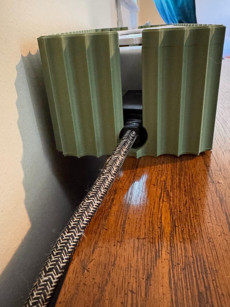 May include: A sage green, 3D-printed cable management box with a vertical ribbed texture. The rectangular enclosure features a circular cutout on the front panel, through which a black and white braided power cord extends outward.