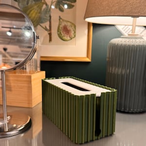 May include: A rectangular tissue box cover featuring a dark olive green exterior with a vertical ribbed texture and a flat white top panel. The cover has a central oval opening for dispensing tissues and a vertical slit on the side for easy access.