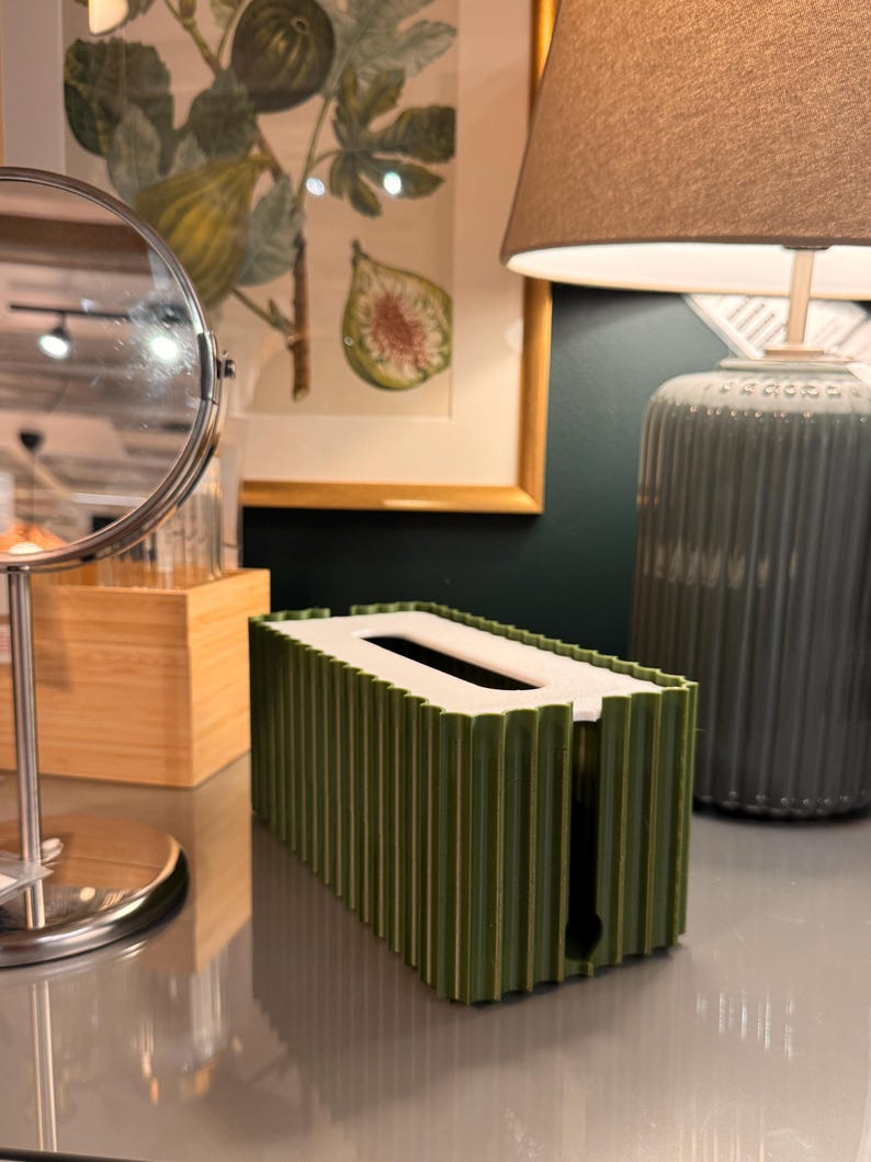 May include: A rectangular tissue box cover featuring a dark olive green exterior with a vertical ribbed texture and a flat white top panel. The cover has a central oval opening for dispensing tissues and a vertical slit on the side for easy access.