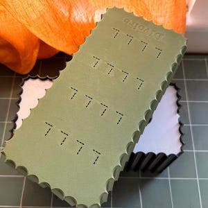 May include: A rectangular olive green box with a scalloped edge and a white interior. The lid features a repeating pattern of small, dashed black lines and the word Divoire embossed on the top surface. The box has a structured, rigid design.