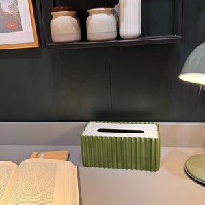May include: A rectangular tissue box cover with a vibrant green, vertically pleated exterior and a smooth white top surface featuring a central elongated opening for dispensing tissues, positioned on a flat surface next to an open book.