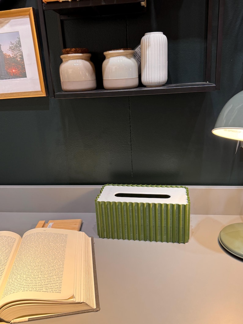 May include: A rectangular tissue box cover with a vibrant green, vertically pleated exterior and a smooth white top surface featuring a central elongated opening for dispensing tissues, positioned on a flat surface next to an open book.