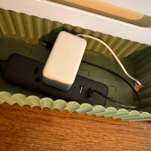 May include: A sage green cable management box with a scalloped, corrugated wall design. Inside, a black power strip with a white power adapter and connected black and white charging cables are neatly organized within the rectangular container.