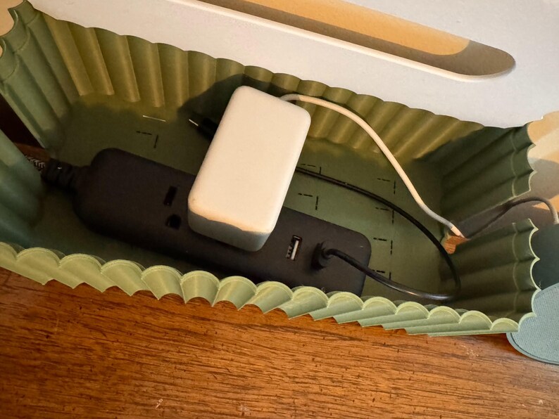 May include: A sage green cable management box with a scalloped, corrugated wall design. Inside, a black power strip with a white power adapter and connected black and white charging cables are neatly organized within the rectangular container.