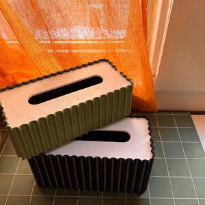 May include: Two rectangular tissue box covers with a scalloped edge design. The top cover is olive green with a white top panel, and the bottom cover is black with a white top panel. Both feature a central elongated oval opening for tissue access.