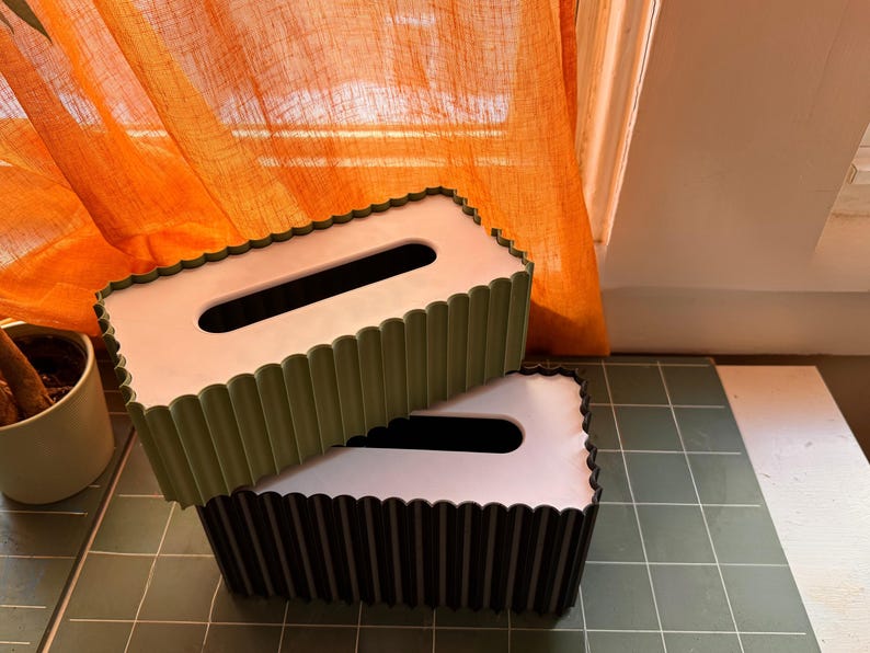 May include: Two rectangular tissue box covers with a scalloped edge design. The top cover is olive green with a white top panel, and the bottom cover is black with a white top panel. Both feature a central elongated oval opening for tissue access.