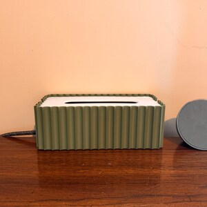 May include: A rectangular olive green tissue box cover with a vertical ribbed texture and a white top panel featuring a central oval opening. Beside the tissue box sits a small, circular, flat gray object with a smooth, solid surface.