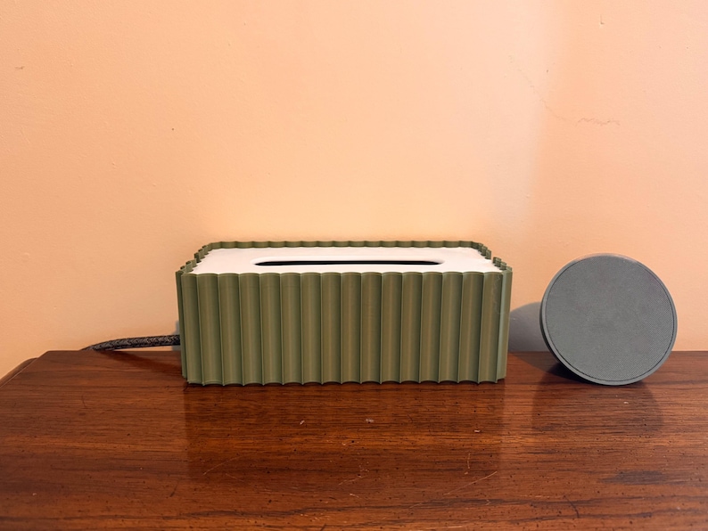 May include: A rectangular olive green tissue box cover with a vertical ribbed texture and a white top panel featuring a central oval opening. Beside the tissue box sits a small, circular, flat gray object with a smooth, solid surface.