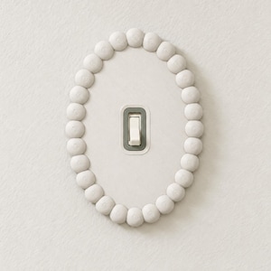 May include: An oval light switch cover plate featuring a textured white finish and a border of small, rounded bead-like elements. The center contains a single white toggle switch set within a rectangular gray frame on a white surface.