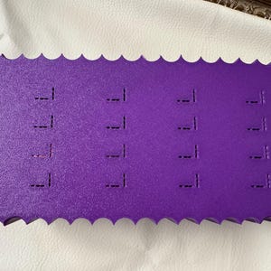 May include: A rectangular purple foam display board with a scalloped edge, featuring four columns of four vertical slits for organizing items. The word "Difoire" is embossed on the right side of the textured surface of this storage accessory.
