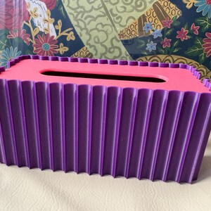 May include: A rectangular tissue box cover featuring a vibrant purple exterior with a vertical fluted or pleated texture. The top surface is a contrasting bright pink color with a centered, elongated oval opening for dispensing tissues.