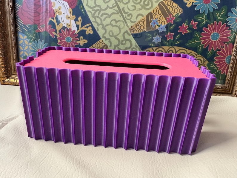 May include: A rectangular tissue box cover featuring a vibrant purple exterior with a vertical fluted or pleated texture. The top surface is a contrasting bright pink color with a centered, elongated oval opening for dispensing tissues.